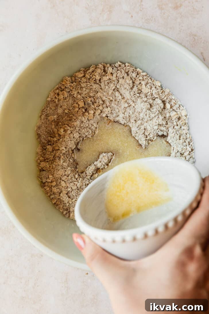 A hand pouring a white bowl of melted butter into a white bowl of flour, oats, sugar, & spices.