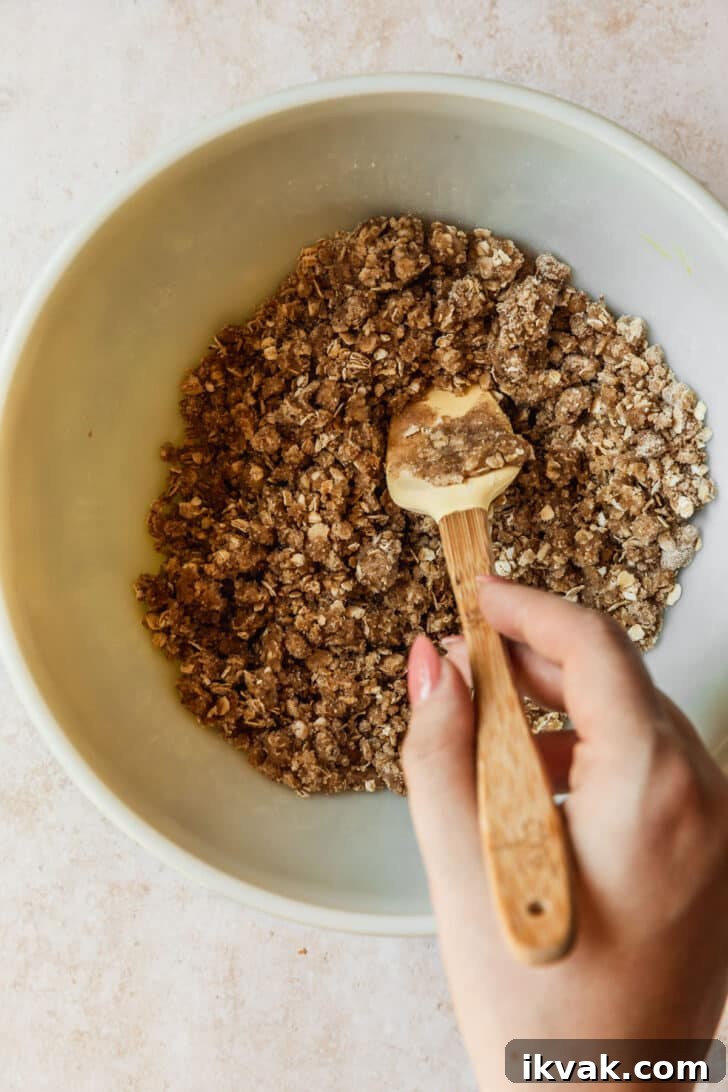 A hand using a spatula to mix oat topping in a white bowl.