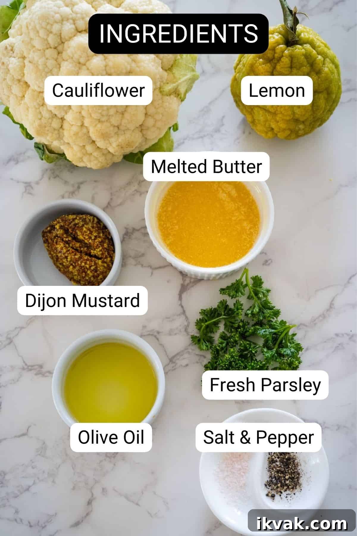 Top-down view of various ingredients labeled for a keto roasted cauliflower recipe, including cauliflower, lemon, melted butter, dijon mustard, olive oil, fresh parsley, and salt & pepper on a marble surface