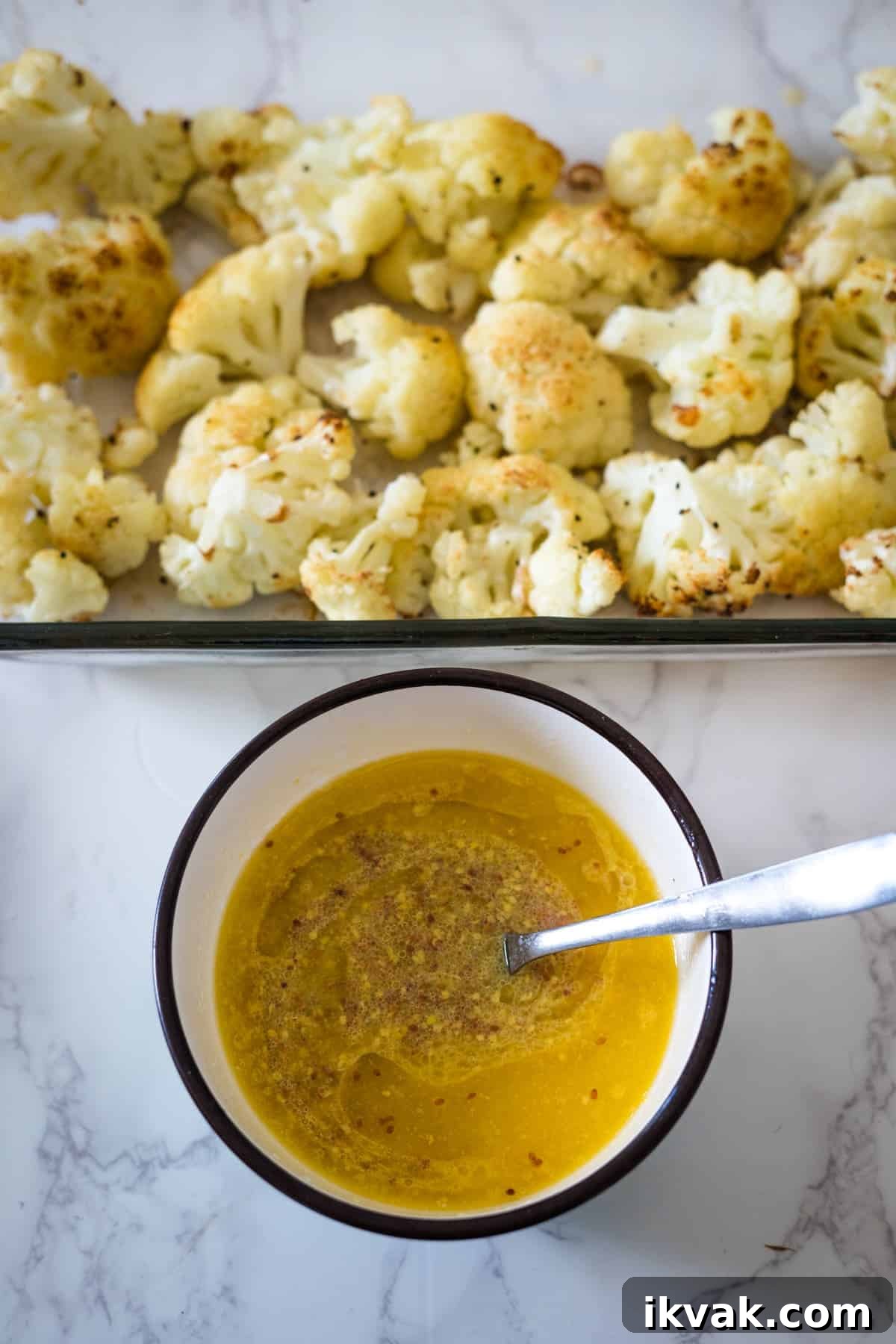 A bowl of yellow sauce with a spoon, next to a tray of keto roasted cauliflower on a marble countertop.