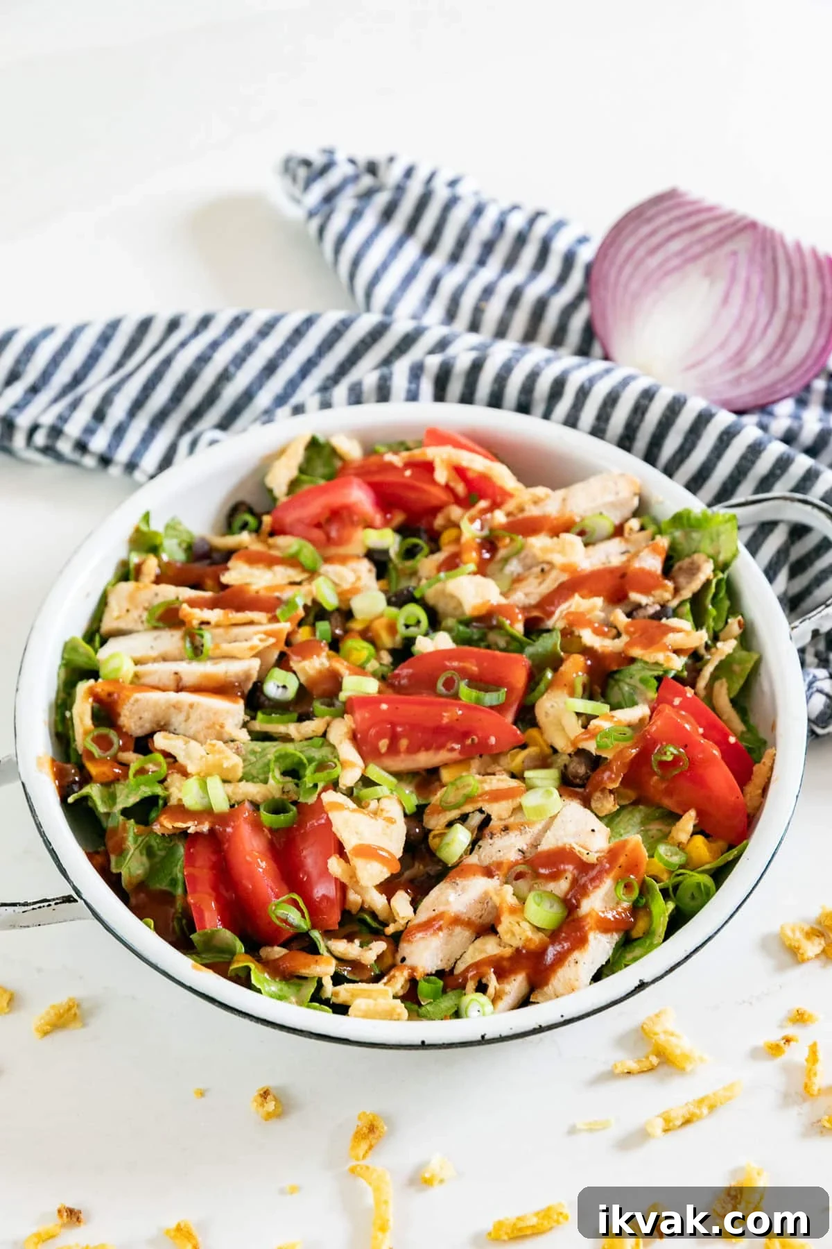 A bowl of BBQ chicken salad with a striped napkin, half red onion and fried onions around it.
