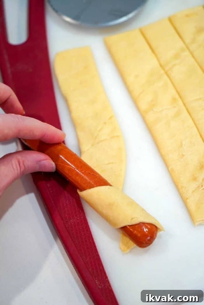 A hand wraps a sausage with a strip of dough on a cutting board, preparing delicious air fryer pigs in a blanket.