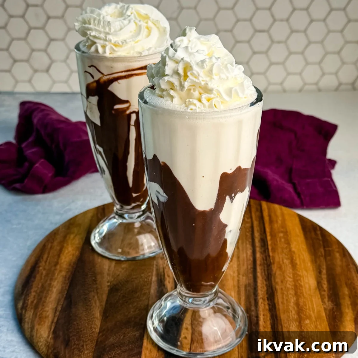 Ninja Slushy Mudslide Recipe: Frozen Coffee Cocktail Guide 2 Two tall glasses filled with creamy Ninja Slushi Mudslides swirled with chocolate syrup and topped with whipped cream, placed on a wooden board with a purple cloth in the background.