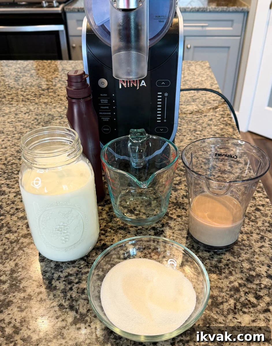 Ninja Slushy Mudslide Recipe: Frozen Coffee Cocktail Guide 3 Ingredients for a Ninja Slushi Mudslide arranged on a countertop, including a jar of milk mixture, a small bowl of sugar, a measuring cup with Irish cream, and chocolate syrup in front of the Ninja Creami machine.