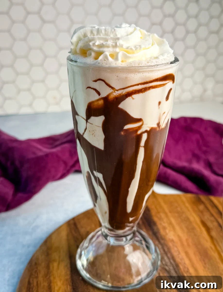 Ninja Slushy Mudslide Recipe: Frozen Coffee Cocktail Guide 8 Close-up of one tall glass filled with a Ninja Slushi Mudslide. The glass is drizzled with chocolate syrup and topped with whipped cream, sitting on a wooden board with a hexagon-tile backsplash in the background.