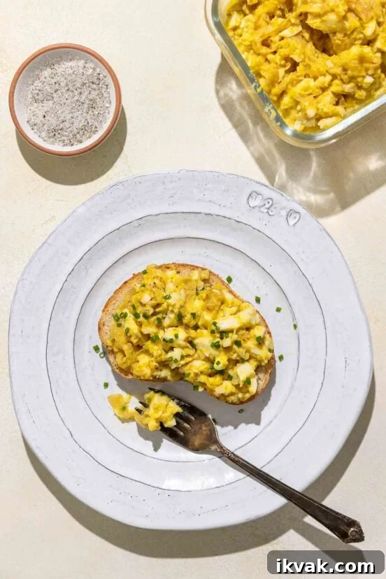 An open-faced egg salad sandwich topped with minced chives on a white plate with a bowl of salad and a dish of salt and pepper nearby.