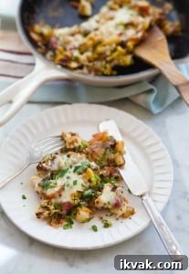 A white plate of fried potatoes with eggs, covered in cheese with a skillet in the background and a fork and knife on the plate.