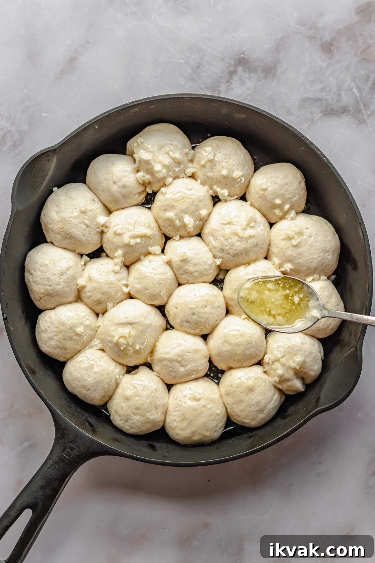 A spoon drizzles melted butter and minced garlic on top of puffy pre-baked bread bites.
