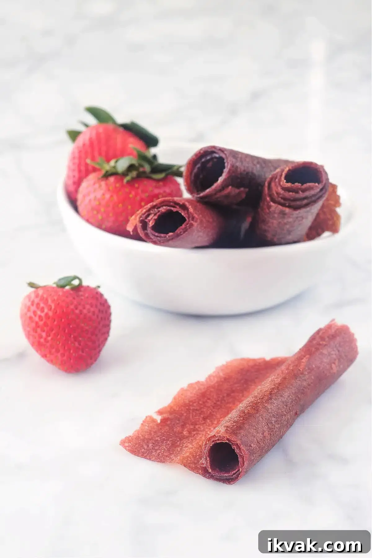 A small white bowl of strawberry basil fruit leather and fresh strawberries. One roll of leather is unrolled and sitting outside the bowl.