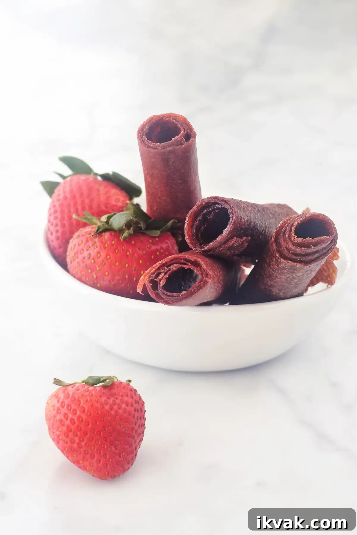 A small white bowl of strawberry basil fruit leather rolled up and sitting in the bowl with fresh strawberries.