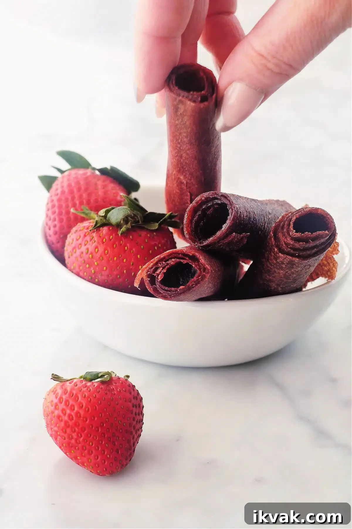 A hand picking up one strawberry fruit roll up from a bowl full of fruit leather and fresh strawberries.
