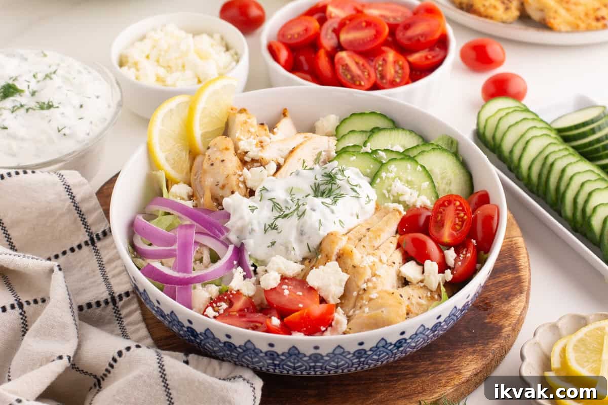 Chicken gyro bowl with grilled chicken slices, halved cherry tomatoes, cucumber slices, red onion, feta, and tzatziki sauce topped with dill. The bowl sits on a wooden board surrounded by small bowls of ingredients and a beige towel with black stripes.