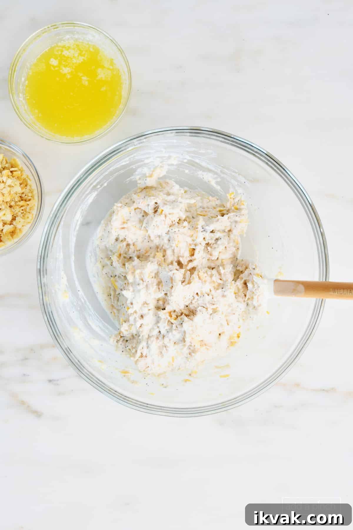 Glass mixing bowl with mixed ingredients for poppy seed chicken casserole. Surrounded by crushed butter crackers and melted butter on a white marble countertop.