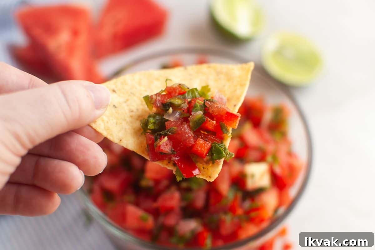 A hand holds a tortilla chip topped with red salsa, with a bowl of salsa, watermelon slices, and halved limes visible in the background.