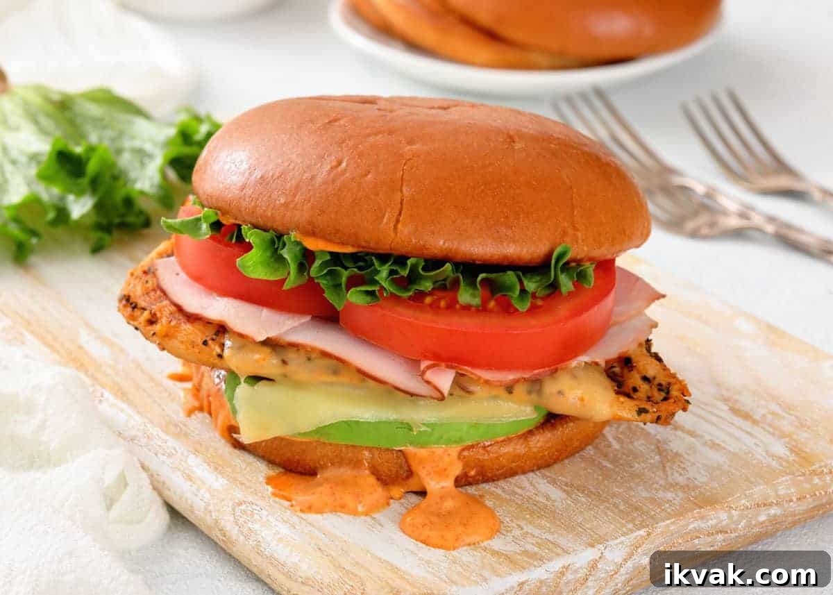 A chicken sandwich with lettuce, tomato, ham, cheese, and sauce on a brioche bun, sitting on a wooden board.
