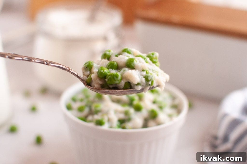 A spoonful of creamed peas is held above a white ramekin filled with the same dish, with green peas visible throughout.
