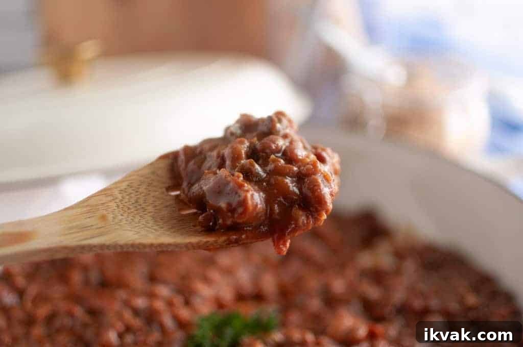 A wooden spoon holds a serving of Baked Bean Casserole above a pot filled with more bean casserole.