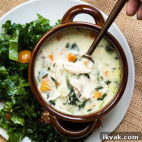 A bowl of creamy chicken Florentine soup with a spoon, served with a side of leafy green salad and cherry tomatoes on a white plate.