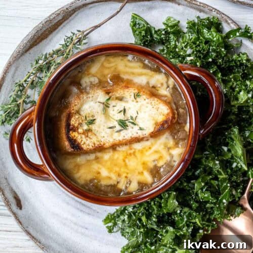 A bowl of French onion soup topped with melted cheese and bread, garnished with herbs. Served on a plate with a side of kale salad and a sprig of thyme.