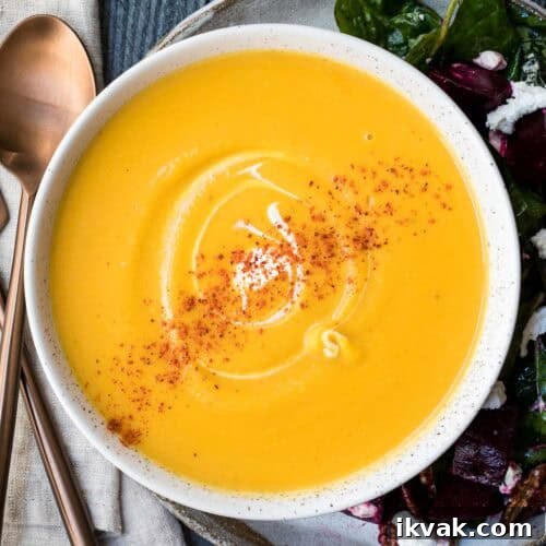 A bowl of creamy butternut squash soup garnished with a swirl of cream and sprinkled with paprika, served next to a salad on a plate with rose gold utensils.
