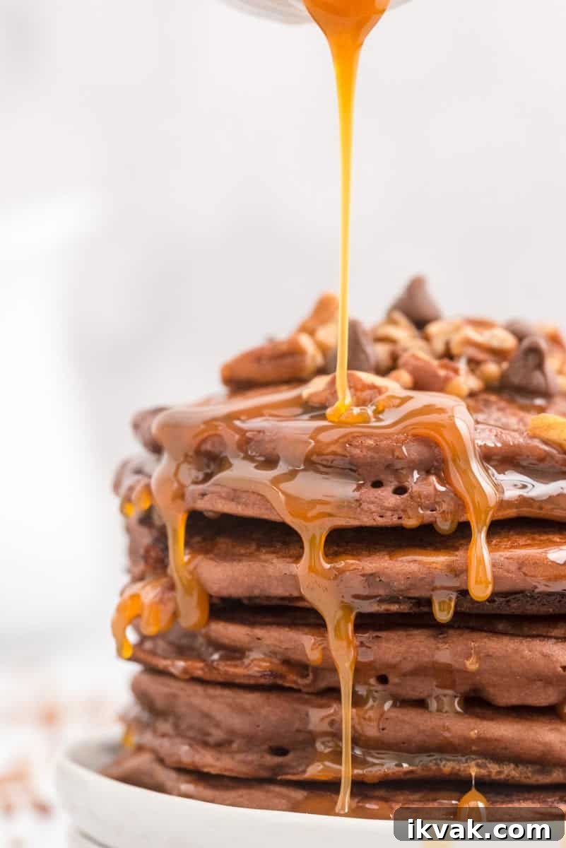 A stack of turtle pancakes with pecans and chocolate chips is topped with caramel being poured over them.