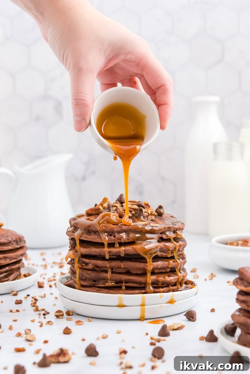 A hand pours syrup over a stack of chocolate pancakes topped with pecan pieces and chocolate chips on a plate.