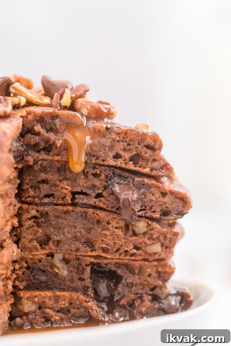 Close-up of a stack of chocolate pancakes with visible pecans and gooey syrup dripping down the sides.