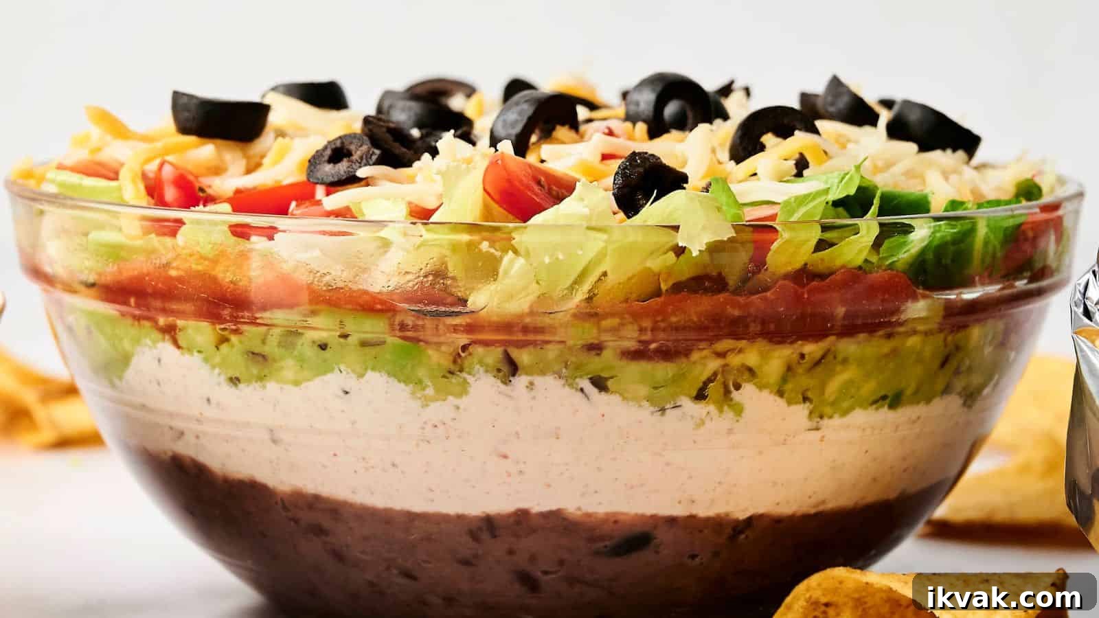 25 Creative Ways to Use Dip Recipes Beyond the Bowl 27 A clear glass bowl with 7 Layer Dip featuring beans, white cream, guacamole, diced tomatoes, shredded cheese, lettuce, and black olives.