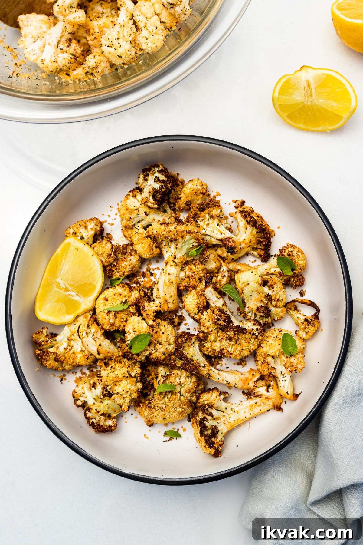 A dish of air fried cauliflower from above with fresh herbs scattered on top and a segment of lemon, with more lemon and a pale blue tea towel.