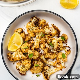 A dish of air fried cauliflower from above with fresh herbs scattered on top and a segment of lemon, with more lemon and a pale blue tea towel.