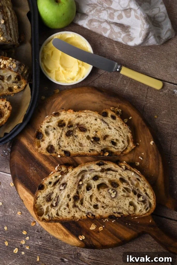 Apple Crisp Sourdough Loaf 2 2 slices of sourdough apple crisp bread laid on a round wooden board so you can see the crumb inside. Around the slices of bread you can see a dish of butter, a floral dish towel and a green apple.