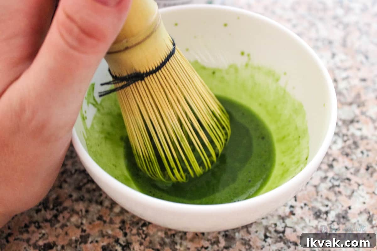 Creamy Strawberry Matcha Smoothie Recipe 3 Hand using a bamboo whisk to mix matcha powder and water in white bowl.