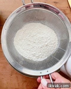 Moist Cherry Cake with Fresh Cherry Glaze 5 Sifting flour mixture over bowl set on a wooden surface.