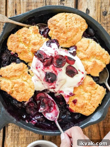 Close up of cast iron skillet with cherry cobbler. Wooden table. Silver spoons.