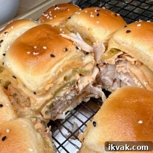 Close-up of golden slider sandwiches filled with shredded chicken, melted cheese, and pickles, topped with sesame seeds, resting on a cooling rack. One slider is pulled away, showing the creamy filling inside.